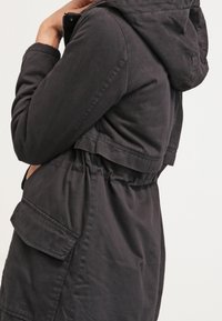 Osoba w ciemnej kurtce z kapturem, z dużą kieszenią i elastycznym pasem, stoi bokiem z ręką przy brodzie.