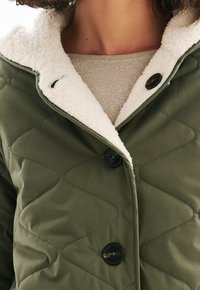 Vihreä tikattu takki, jossa on suuret napit ja valkoinen sherpa fleece-vuori, päällä beige kiiltävä paita, näkyvissä rintakehältä ylöspäin.