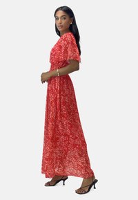 Red floral maxi dress with short, sheer sleeves, lightweight fabric, and a flowing silhouette. Black heeled sandals complete the look.