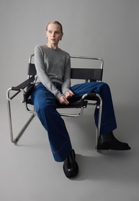 Gray knitted sweater, blue wide-leg jeans with red stitching, and black ankle boots. Seated on a modern black and chrome chair.