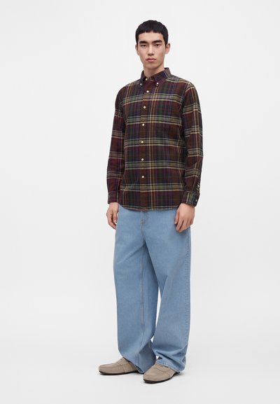 Jeune homme debout portant une chemise à carreaux foncée boutonnée, un jean bleu clair ample et des mocassins beiges sur un fond blanc uni.