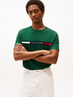 Zelené bavlněné tričko s krátkým rukávem, které má horizontální pruhovaný design v bílé, červené a námořnické, s textem "TOMMY HILFIGER" uprostřed.