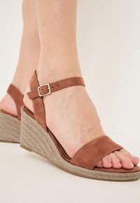 Sandales compensées en daim marron avec une semelle en corde texturée, dotées d'une bride de cheville avec une boucle dorée et d'un design à bout ouvert.