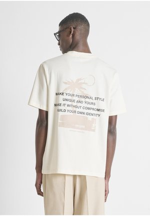 Homme portant un T-shirt écru avec un texte noir et un motif de palmier dans le dos, assorti à un pantalon beige et des lunettes.