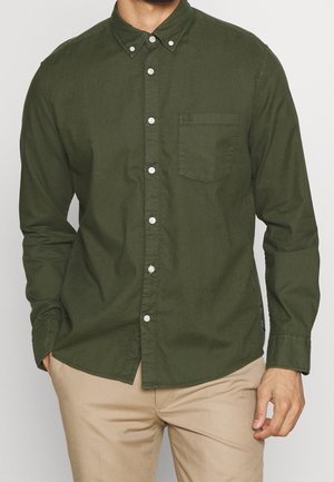 Mann trägt ein olivgrünes Hemd mit Knopfleiste und Taschen auf der Vorderseite sowie eine beige Hose und steht mit einer Hand in der Tasche.