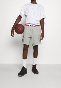 Szare spodenki z czerwono-niebieskim paskiem i logo NBA, zestawione z białą koszulką z małymi logo NBA. Czarne trampki i białe skarpetki.