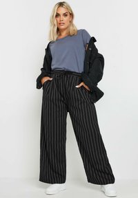 Pantaloni neri a gamba larga con strisce verticali bianche, vita elasticizzata con cordoncino, abbinati a una t-shirt grigia e una giacca nera. Sneakers bianche.