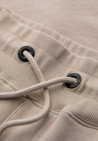 Gros plan sur la ceinture de pantalon de survêtement beige avec des côtes cousues, deux œillets en métal noir et un cordon tressé assorti passé à travers eux.