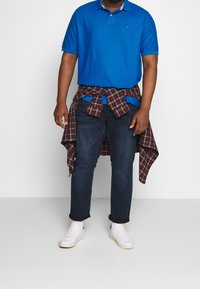 Blå pikétröja med krage, kombinerad med mörka jeans. En röd och grå rutigt skjorta är knuten runt midjan. Vita sneakers på fötterna.