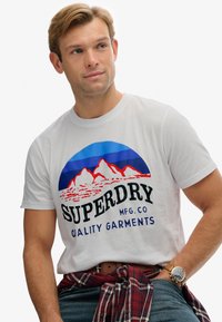 Hvid bomulds T-shirt med et farverigt bjergmotiv i blå, rød og hvid. Sort tekst læser "SUPERDRY MFG. CO KVALITETSTØJ."
