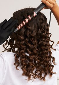 Black curling iron with a pink spiral barrel in use on dark brown, curly hair. User wears a black heat-resistant glove. White shirt visible.