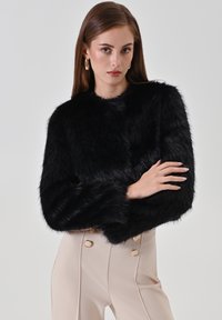 Veste courte en fausse fourrure noire avec manches longues, dotée d'une texture douce et d'un col arrondi, associée à un pantalon beige taille haute.