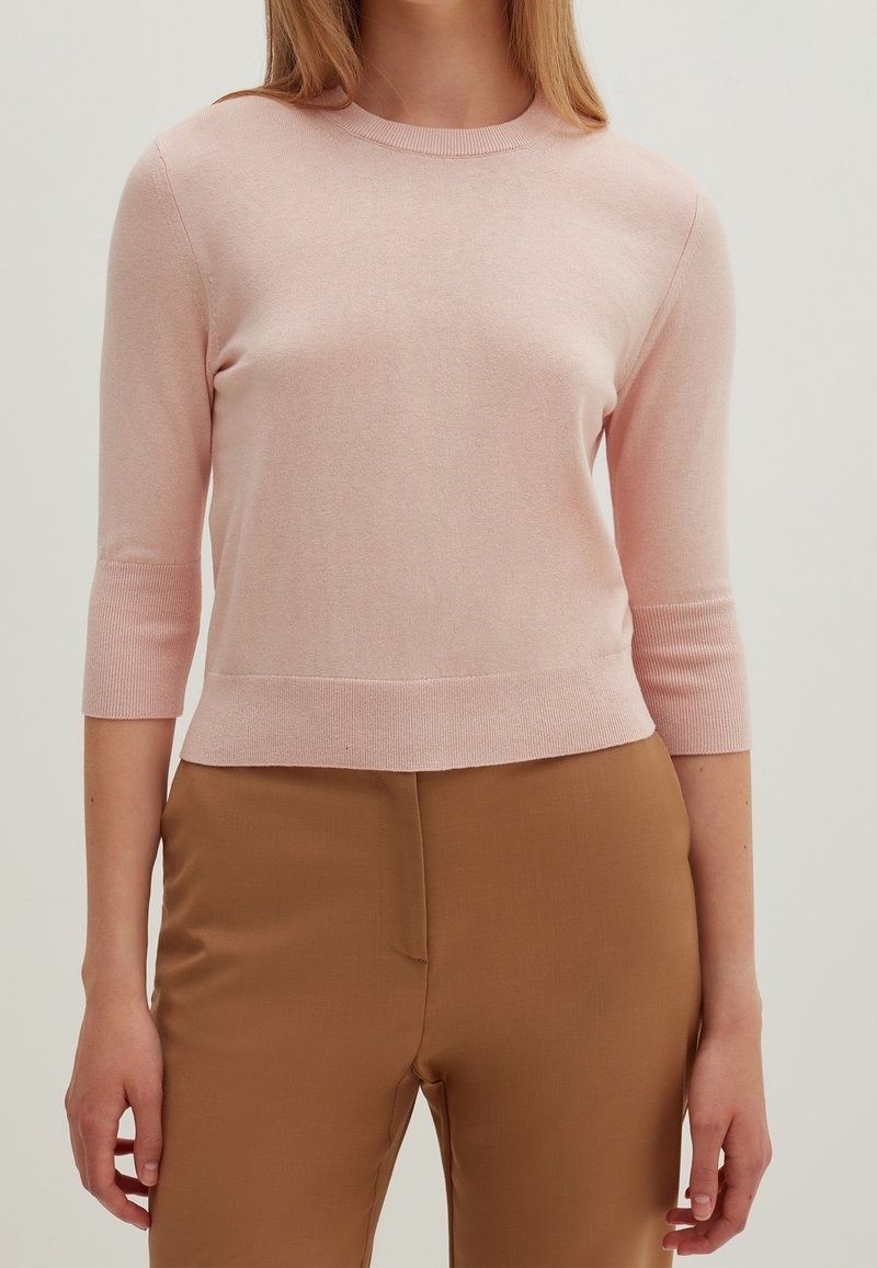 Jasnoróżowy dopasowany sweter z rękawami do połowy przedramienia, prążkowanym dekoltem i mankietami, zestawiony z beżowymi spodniami. Widoczna miękka, dzianinowa faktura.