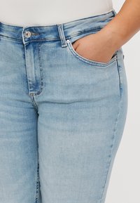 Svijetloplave traperice ravnog kroja, s prednjim dzepom, metalnim gumbom na stražnjem dijelu i suptilnim izbljeđivanjem na tkanini.