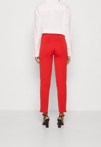Pantalon rouge ajusté en matière extensible, associé à une chemise à col rose clair. Le pantalon présente une texture lisse et une coupe slim.