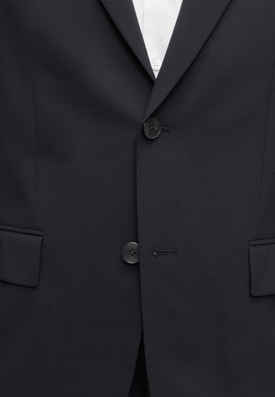 Gros plan d'une veste de costume noire boutonnée sur une chemise blanche, montrant deux boutons et des poches à rabat de chaque côté.