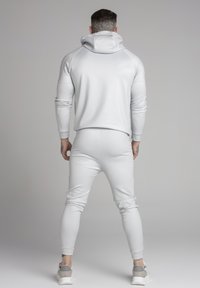 Tuta sportiva grigio chiaro con cappuccio, design aderente, tessuto elasticizzato, polsini a coste e pantaloni tapered abbinati. Calzature casual visibili.