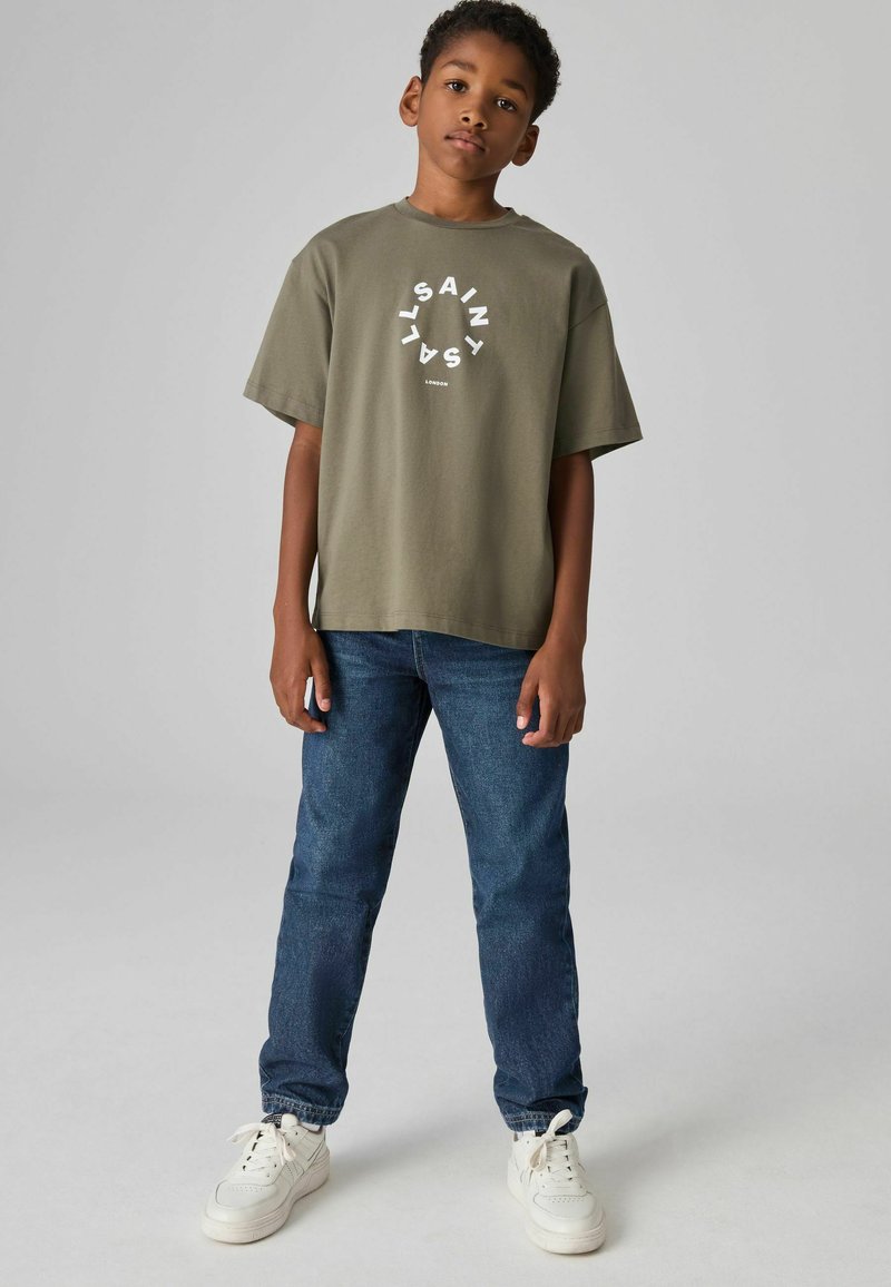 Camiseta de algodón verde oliva con texto gráfico circular blanco, combinada con jeans de mezclilla azules y zapatillas blancas. Ajuste simple y relajado.
