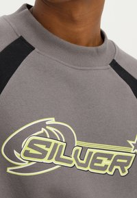 Felpa grigia con accenti neri sulle spalle, caratterizzata da un vivace testo grafico giallo e nero "SILVER" e elementi decorativi.