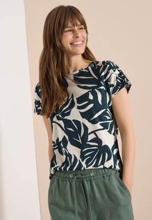 Jeune femme souriante, portant une chemise beige à manches courtes avec un imprimé de feuilles tropicales bleu foncé et un pantalon vert à cordon, debout les mains dans les poches.