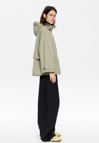 Giacca impermeabile verde chiaro con cappuccio, taglio loose e tasca frontale. Abbinata a pantaloni neri a gamba larga e scarpe chiare.