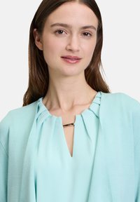 Lichtblauwe cardigan met een zachte textuur, voorzien van een geplooide halslijn en een sluiting in goudkleur. Glad materiaal en een comfortabele pasvorm.