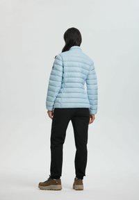 Giacca a vento azzurra chiara con cuciture orizzontali, colletto alto e polsini elasticizzati, abbinata a pantaloni neri e scarponcini da trekking grigi.