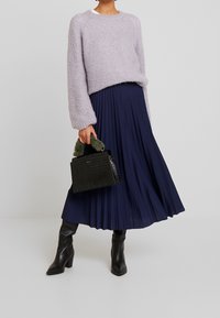 Maglione lavorato a maglia viola chiaro, gonna a pieghe blu scuro, stivali neri fino al ginocchio e una piccola borsa nera in pelle di coccodrillo con dettagli verdi.