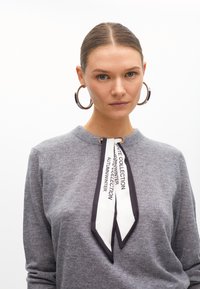 Szary sweter z okrągłym dekoltem, z czarno-białym, wzorzystym szalem zawiązanym z przodu. Modelka nosi duże, srebrne kolczyki w kształcie kół.