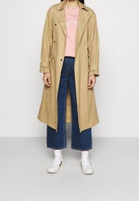 Trench beige con botones de doble botonadura, mangas enrolladas, usado sobre una camisa rosa, combinado con jeans oscuros de pierna ancha y zapatillas blancas.