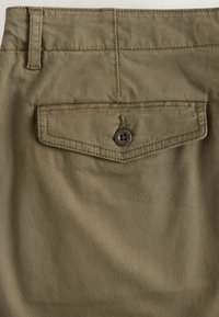 Shorts en tissu kaki avec une poche arrière dotée d'une fermeture à bouton. La texture est lisse avec de légères lignes verticales.