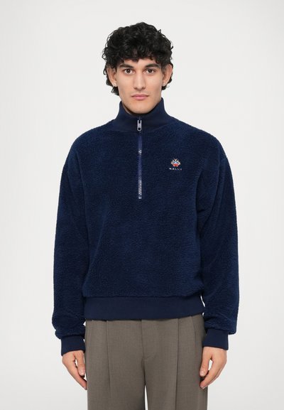 Pull-over en polaire bleu marine avec un col zippé à mi-hauteur, arborant un logo discret. Le tissu présente une finition douce et texturée, avec des poignets et une taille côtelés.