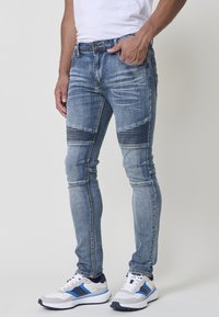 Jeans ajustados de denim azul con detalles de rasgado y desvanecido, que presentan costuras horizontales en las rodillas; combinados con zapatillas deportivas blancas y azules.