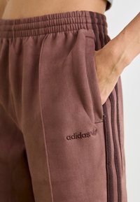 Hnědé sportovní tepláky s texturovaným povrchem, elastickým pasem, bočními kapsami a tónovaným logem Adidas potištěným na levém stehně.