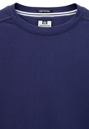 Granatowa bluza z okrągłym dekoltem, z czarnymi i białymi metkami wewnątrz kołnierza z napisem "ADMIT NOTHING" i "WEEKEND" oraz ikoną korony.