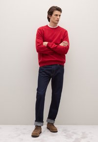 Roter Pullover mit rundem Ausschnitt und gerippten Bündchen, kombiniert mit dunklen Denim-Jeans mit umgeschlagenen Säumen und braunen Schnürstiefeln.
