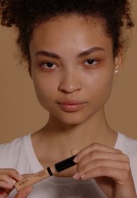 KIKO Full Coverage Liquid Concealer in einer beigen Tube mit schwarzem Deckel, gehalten von einem Model. Ein neutraler Hintergrund hebt die Produkteigenschaften hervor.