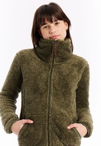 Mladá žena v olivově zelené fleecové bundě se zipem a vysokým límcem, s rukama v kapsách, dívající se na kameru na bílém pozadí.