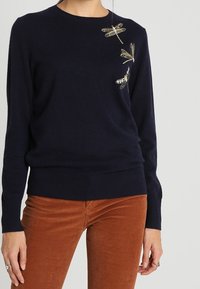 Siniasestä kudottu neuletakki, jossa on pyöreä kaula-aukko, ja vasemmassa olkapäässä kultaisia brodeerattuja sudenkorentokuvioita sekä ribbauslahkeet.