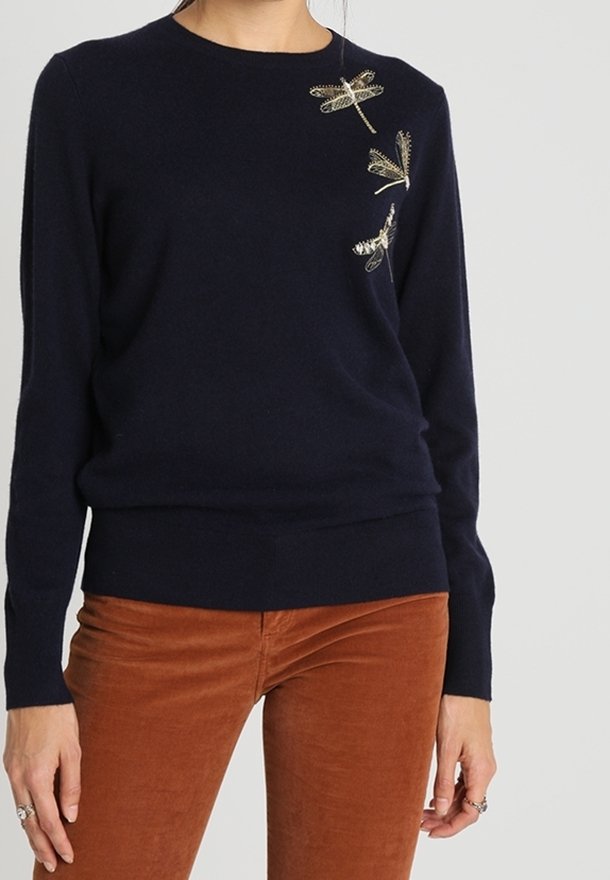 Siniasestä kudottu neuletakki, jossa on pyöreä kaula-aukko, ja vasemmassa olkapäässä kultaisia brodeerattuja sudenkorentokuvioita sekä ribbauslahkeet.