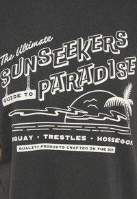 Mustast puuvillasest T-särgist, millel on valge graafiline trükk stiliseeritud tekstist ning rannamaastik palmipuude ja mägedega.