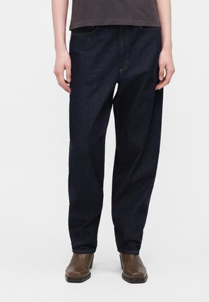 Personne portant un jean bleu foncé ample, des bottines marron et un haut foncé, debout sur fond blanc.