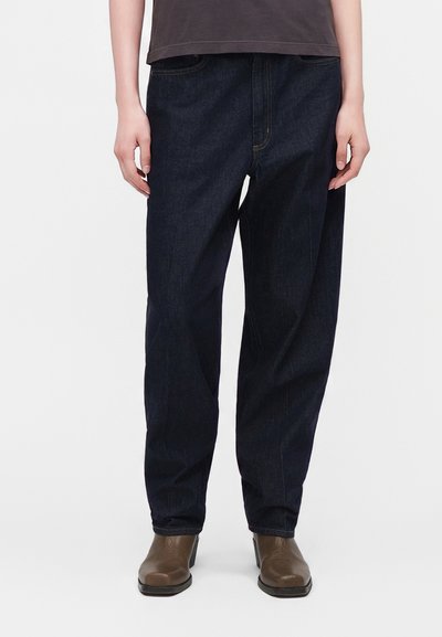 Personne portant un jean bleu foncé ample, des bottines marron et un haut foncé, debout sur fond blanc.