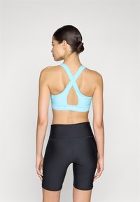 Sujetador deportivo de color azul claro con tirantes cruzados en la parte posterior y una banda con logo, combinado con pantalones cortos de ciclismo negros de cintura alta. Material suave y elástico.