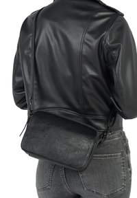 Sac à bandoulière synthétique noir avec une surface texturée, fermeture à glissière et sangle réglable, porté sur une veste en cuir noir.