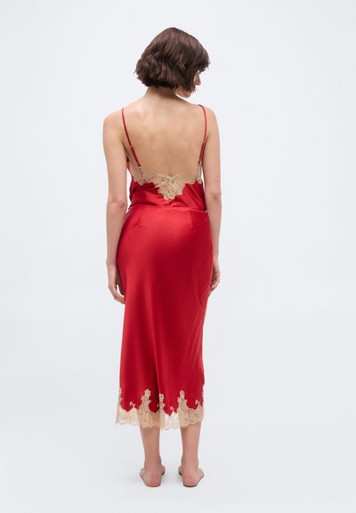 Vrouw die blootsvoets staat met de rug naar voren, gekleed in een rood satijnen slipjurkje met beige kanten rand aan de achterkant en zoom.