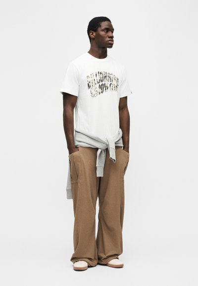 Νεαρός άνδρας που στέκεται φορώντας λευκό μπλουζάκι με σχέδιο, μπεζ παντελόνι φαρδύ, λευκά παπούτσια και γκρι φούτερ δεμένο γύρω από τη μέση του.