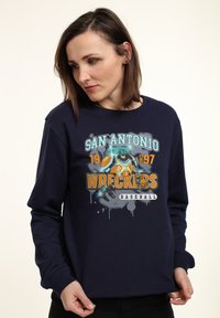 Sudadera azul marino con un estampado gráfico que incluye "San Antonio Wreckers" y una ilustración de un personaje de béisbol. Cuello clásico estilo crew.