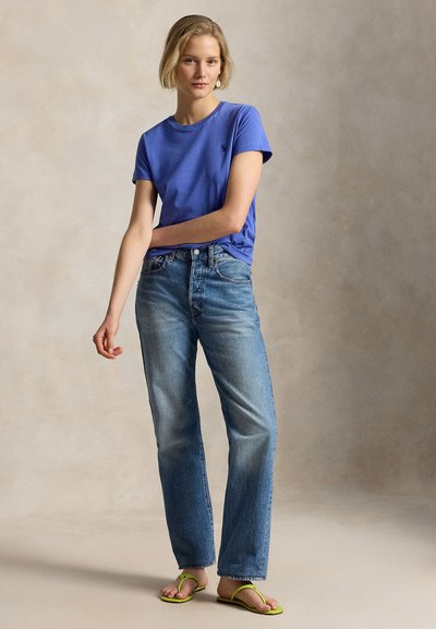 Γυναίκα με κοντά ξανθά μαλλιά, που φοράει μπλε μπλουζάκι, ανοιχτόχρωμο τζιν και πράσινα σαγιονάρες, στέκεται μπροστά σε ουδέτερο φόντο.