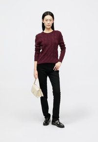 Maglione bordeaux a treccia, pantaloni neri slim-fit, mocassini neri e borsa beige. Modella posa su uno sfondo neutro.